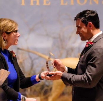 A woman hands a glass trophy to an appreciative man, with a stuffed lion in the background. 