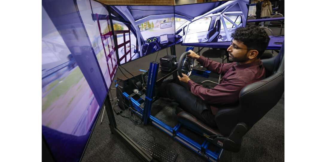 A man sits in a simulated driving cockpit holding a steering wheel surrounded by screens showing a racing video game.