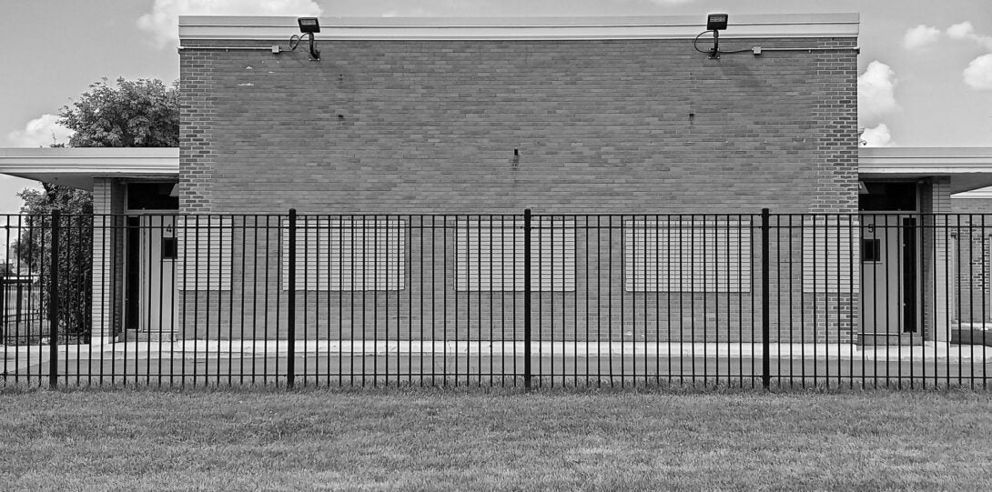 A black and white photograph of a brick building behind a wrought iron fence.