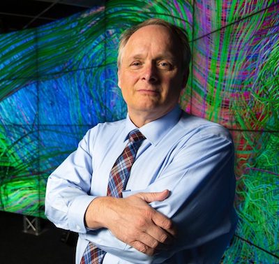 A man in a tie stands in front of a wall of screens showing a scientific image.