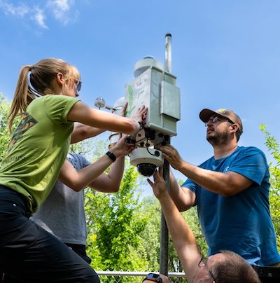 Three scientists installing a gray sensor box attached to a pole.