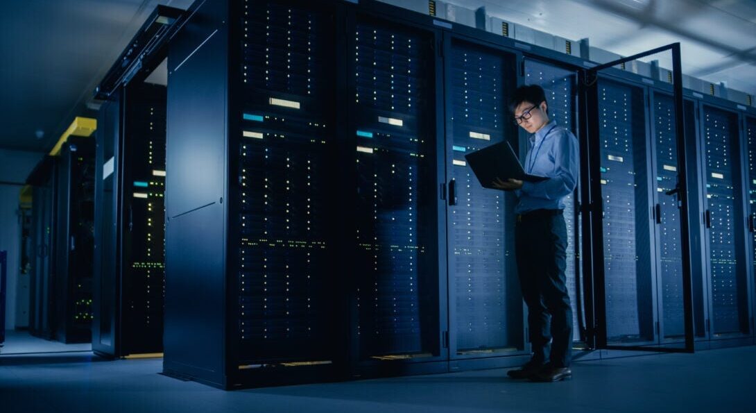 A man looks at a laptop computer in front of a bank of computer servers.