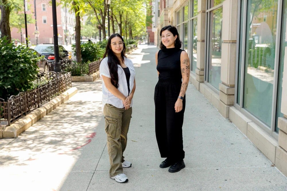 Two women stand on a sidewalk smiling.