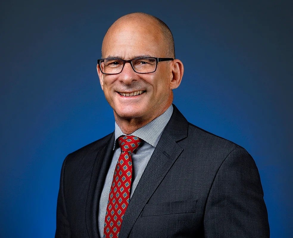 A headshot of a man wearing glasses, a red tie and a black suit against a blue background.