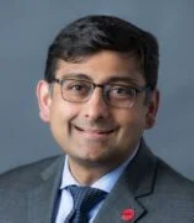 A headshot of a man smiling and wearing glasses and a gray suit and navy tie.