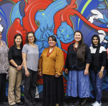 Seven women stand smiling in front of a red, blue and white heart mural.
                  