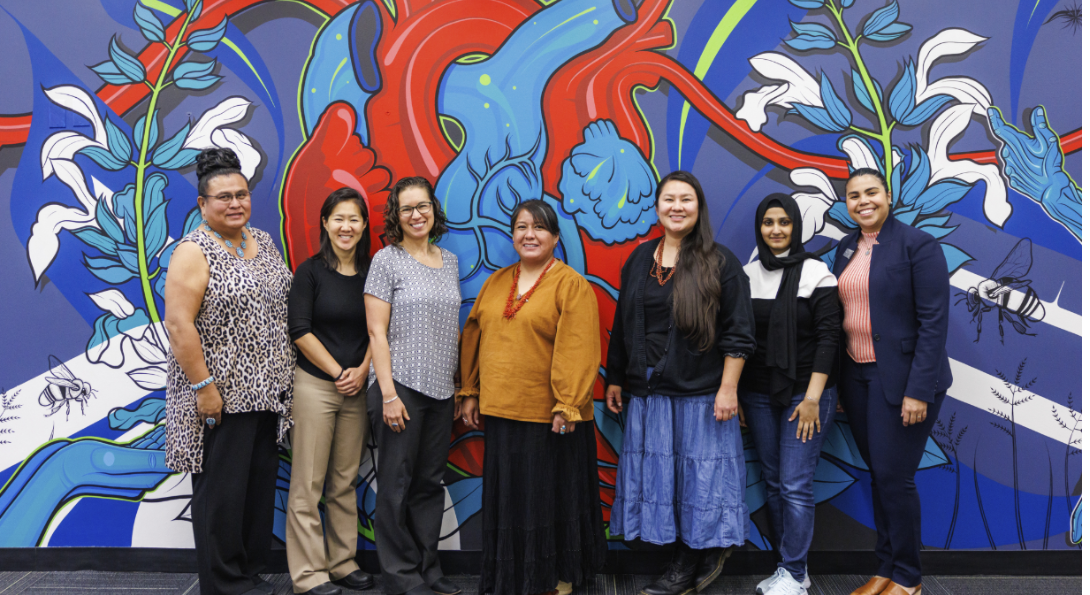 Seven women stand smiling in front of a red, blue and white heart mural.