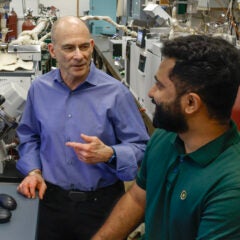 A man in a blue dress shirt points to the left while talking to a bearded man in a green polo.