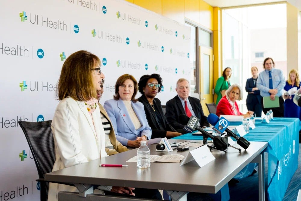 A panel of researchers speak into news microphones in front of a UI Health banner.