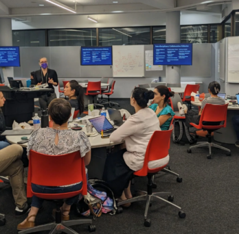 UIC researchers sit at tables in groups of five while listening to a presenter.
                  