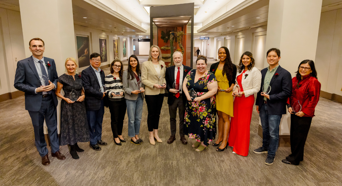 Twelve people stand in a line holding glass awards.